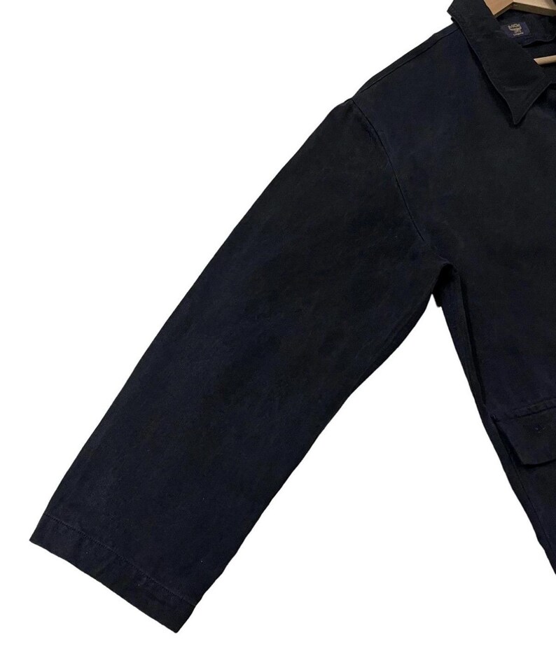 May include: A close-up of a dark navy blue jacket. The jacket has a collar, long sleeves, and a pocket. The fabric appears to be a sturdy material, possibly denim or canvas. The jacket is hanging on a wooden hanger.