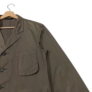 May include: A khaki-colored jacket with a classic collar, three buttons, and a single chest pocket. The jacket is hanging on a wooden hanger against a white background. The jacket's design is simple and functional.