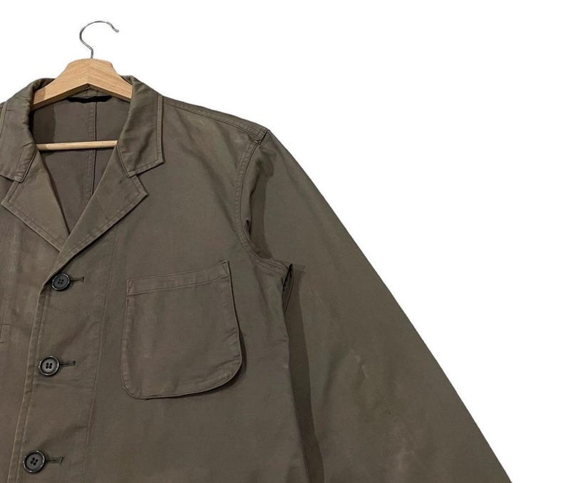 May include: A khaki-colored jacket with a classic collar, three buttons, and a single chest pocket. The jacket is hanging on a wooden hanger against a white background. The jacket's design is simple and functional.