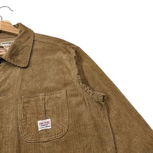 K&ouml;nnte beinhalten: Eine braune Cordjacke mit Knopfleiste und Kragen. Die Jacke hat eine Brusttasche mit einem kleinen rechteckigen Etikett mit der Aufschrift "SOMETHING". Die Jacke h&auml;ngt an einem Holzb&uuml;gel.