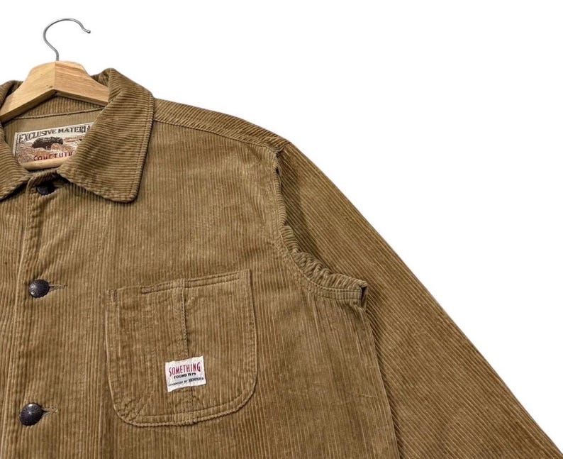 K&ouml;nnte beinhalten: Eine braune Cordjacke mit Knopfleiste und Kragen. Die Jacke hat eine Brusttasche mit einem kleinen rechteckigen Etikett mit der Aufschrift "SOMETHING". Die Jacke h&auml;ngt an einem Holzb&uuml;gel.