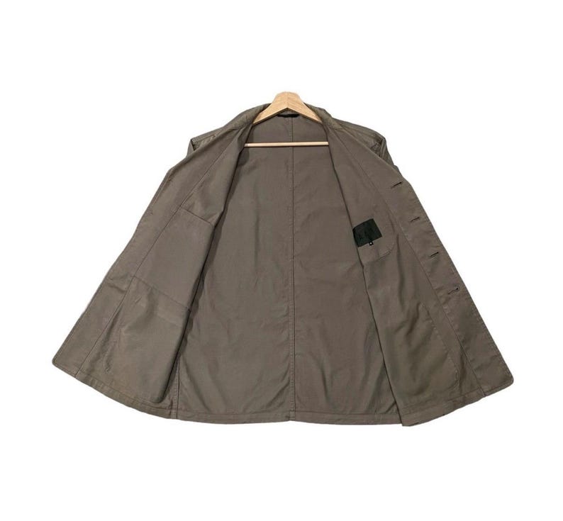 May include: A khaki-colored jacket is displayed open, revealing the interior lining and pockets. The jacket is hanging on a wooden hanger against a white background. The jacket has a classic design with a collar and button closures.