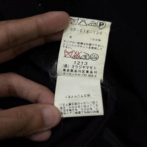 May include: A close-up of a black garment tag held by a hand. The tag has Japanese text and symbols, including washing instructions and the text "HP-C16-130". The tag also includes the text "100%" and the number "1213".