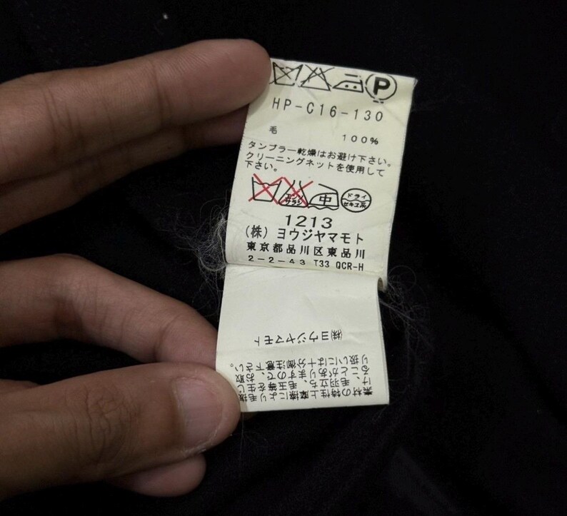May include: A close-up of a black garment tag held by a hand. The tag has Japanese text and symbols, including washing instructions and the text "HP-C16-130". The tag also includes the text "100%" and the number "1213".