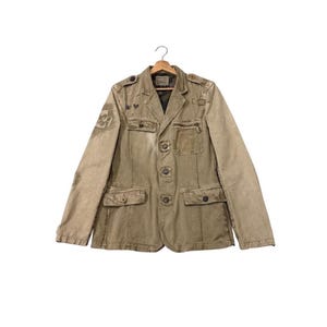 May include: A tan military-style jacket with multiple pockets, buttons, and decorative patches. The jacket is buttoned up and hanging on a wooden hanger. The jacket has a worn, vintage appearance.