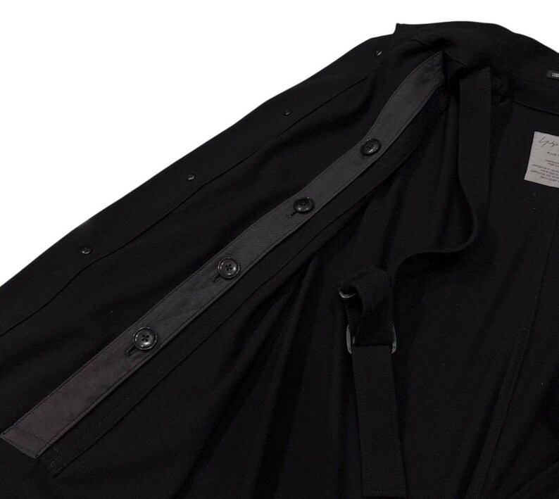 May include: Black jacket with a button-up closure and a strap detail. The jacket features a dark gray strip with buttons and a label on the inside. The fabric appears to be a lightweight material.