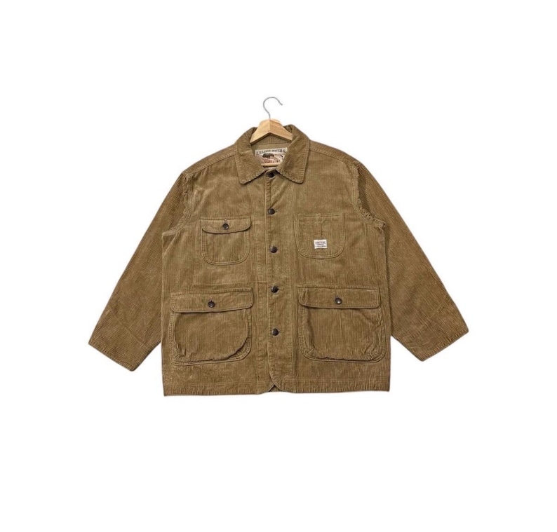 K&ouml;nnte beinhalten: Eine braune Cordjacke mit Kragen und vier Vordertaschen. Die Jacke hat einen Knopfverschluss und ein kleines Etikett auf der rechten Tasche. Die Jacke h&auml;ngt an einem Holzb&uuml;gel.