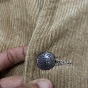 K&ouml;nnte beinhalten: Nahaufnahme einer beigefarbenen Cordjacke. Die Jacke hat einen Knopf mit dem Text "Something" um ein Kronenmuster. Der Cordstoff hat vertikale Rippen.