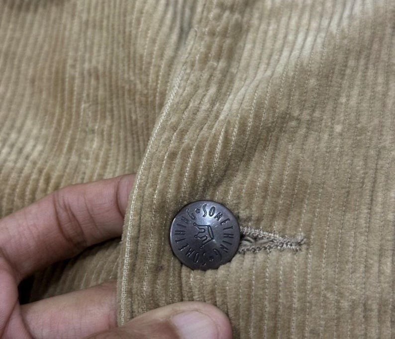 K&ouml;nnte beinhalten: Nahaufnahme einer beigefarbenen Cordjacke. Die Jacke hat einen Knopf mit dem Text "Something" um ein Kronenmuster. Der Cordstoff hat vertikale Rippen.