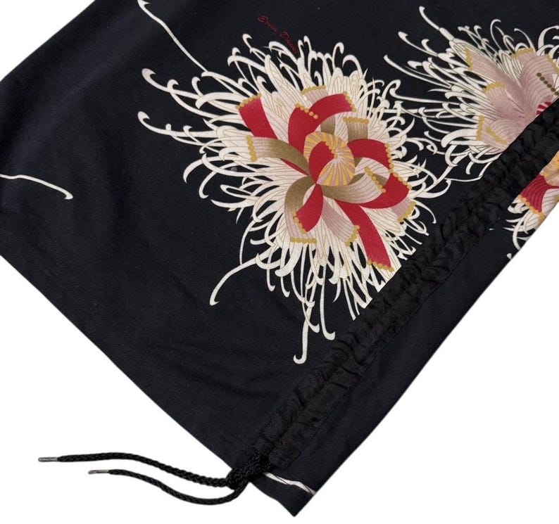May include: Black fabric with a floral design featuring red, white, and gold accents. The design includes a drawstring closure with black cord and metal aglets. The fabric is folded, showing the pattern.