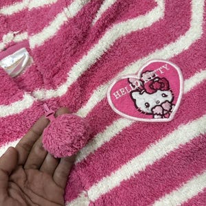 May include: Pink and white striped hooded towel with a Hello Kitty heart-shaped patch. The towel has a pink pom-pom zipper pull. The Hello Kitty patch features the character's face and the words "Hello Kitty".