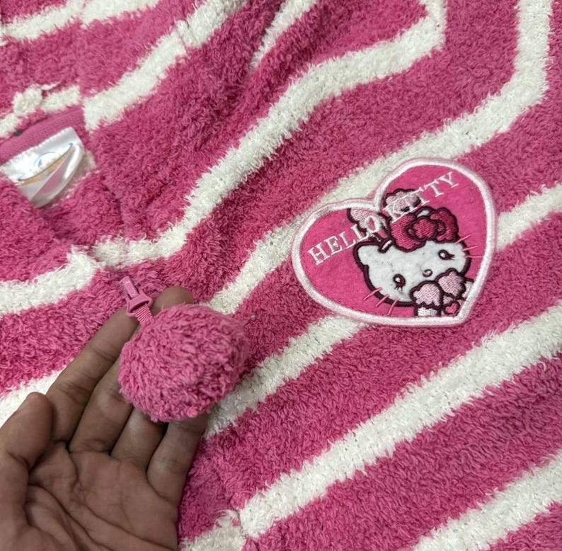 May include: Pink and white striped hooded towel with a Hello Kitty heart-shaped patch. The towel has a pink pom-pom zipper pull. The Hello Kitty patch features the character's face and the words "Hello Kitty".
