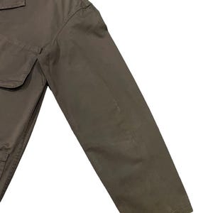 May include: A close-up of a khaki-colored jacket with a collar and two front pockets. The jacket's sleeve is angled towards the bottom right corner, showcasing the fabric's texture and color.