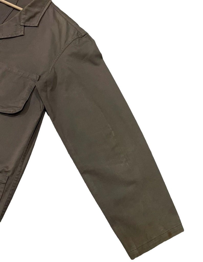 May include: A close-up of a khaki-colored jacket with a collar and two front pockets. The jacket's sleeve is angled towards the bottom right corner, showcasing the fabric's texture and color.