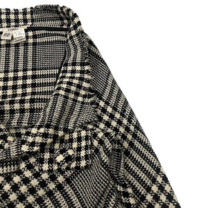 May include: Close-up of a black and white houndstooth patterned jacket. The fabric has a textured appearance. The jacket has a collar and a label is visible.