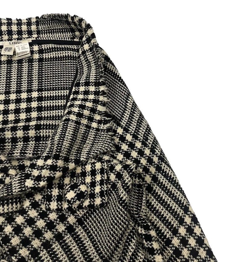 May include: Close-up of a black and white houndstooth patterned jacket. The fabric has a textured appearance. The jacket has a collar and a label is visible.