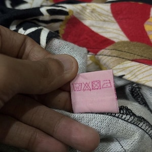 May include: A pink clothing label with care instructions is held against a grey sweatshirt. The background shows a colourful floral pattern with red, white, and black elements. The label has various care symbols.