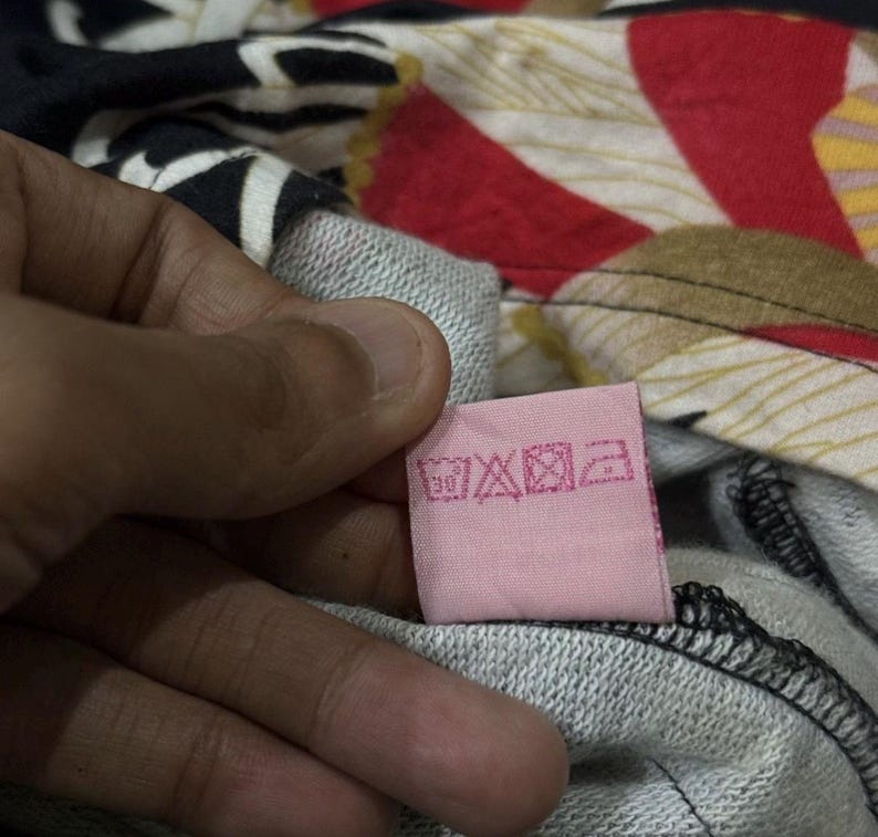 May include: A pink clothing label with care instructions is held against a grey sweatshirt. The background shows a colourful floral pattern with red, white, and black elements. The label has various care symbols.