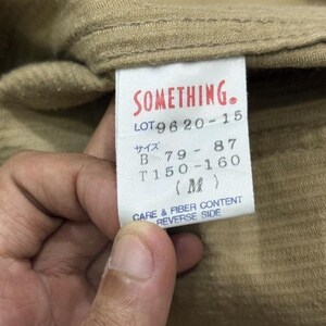 K&ouml;nnte beinhalten: Nahaufnahme eines beigefarbenen Stoffes mit einem wei&szlig;en Etikett. Das Etikett hat roten Text mit der Aufschrift "SOMETHING." Darunter befinden sich Zahlen und japanische Schriftzeichen. Das Etikett lautet auch "CARE & FIBER CONTENT REVERSE SIDE."