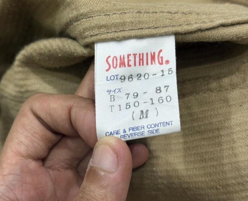 K&ouml;nnte beinhalten: Nahaufnahme eines beigefarbenen Stoffes mit einem wei&szlig;en Etikett. Das Etikett hat roten Text mit der Aufschrift "SOMETHING." Darunter befinden sich Zahlen und japanische Schriftzeichen. Das Etikett lautet auch "CARE & FIBER CONTENT REVERSE SIDE."