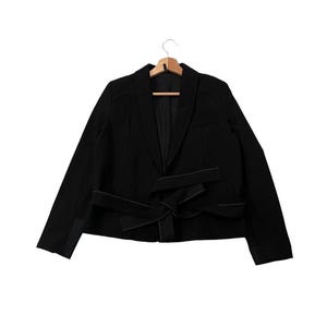 May include: A black blazer with a notched lapel and a unique tie detail at the waist. The blazer is hanging on a wooden hanger. The sleeves are long and the overall design is modern and stylish.