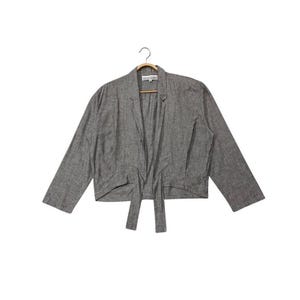 May include: A gray blazer with long sleeves, hanging on a wooden hanger. The blazer has a tie closure at the waist and a notched collar. The fabric appears to be a textured linen or similar material.