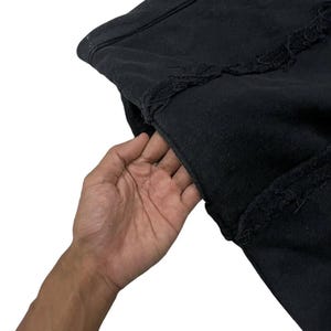 Puede incluir: Primer plano de una prenda negra con bordes deshilachados. La tela parece ser un material pesado, posiblemente una sudadera o chaqueta. Una mano se mete en un bolsillo.
