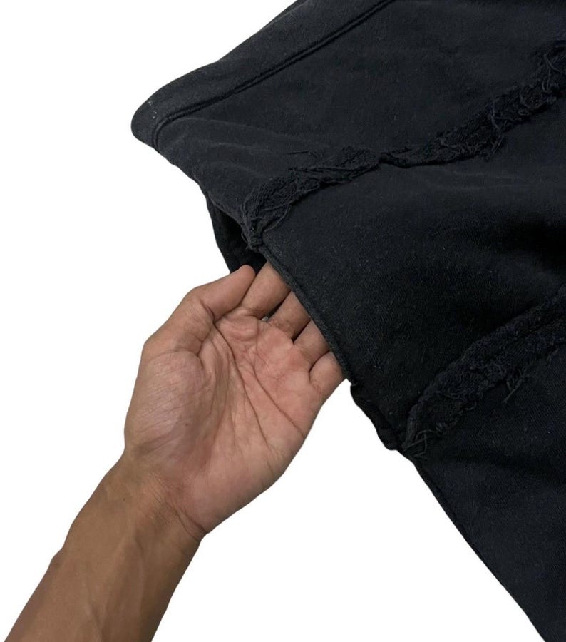 Puede incluir: Primer plano de una prenda negra con bordes deshilachados. La tela parece ser un material pesado, posiblemente una sudadera o chaqueta. Una mano se mete en un bolsillo.