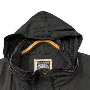 Puede incluir: Chaqueta con capucha negra con cierre de cremallera y percha de madera. La chaqueta presenta una peque&ntilde;a etiqueta con el texto "Von Dutch" y un cierre de bot&oacute;n. La chaqueta es de tela oscura.