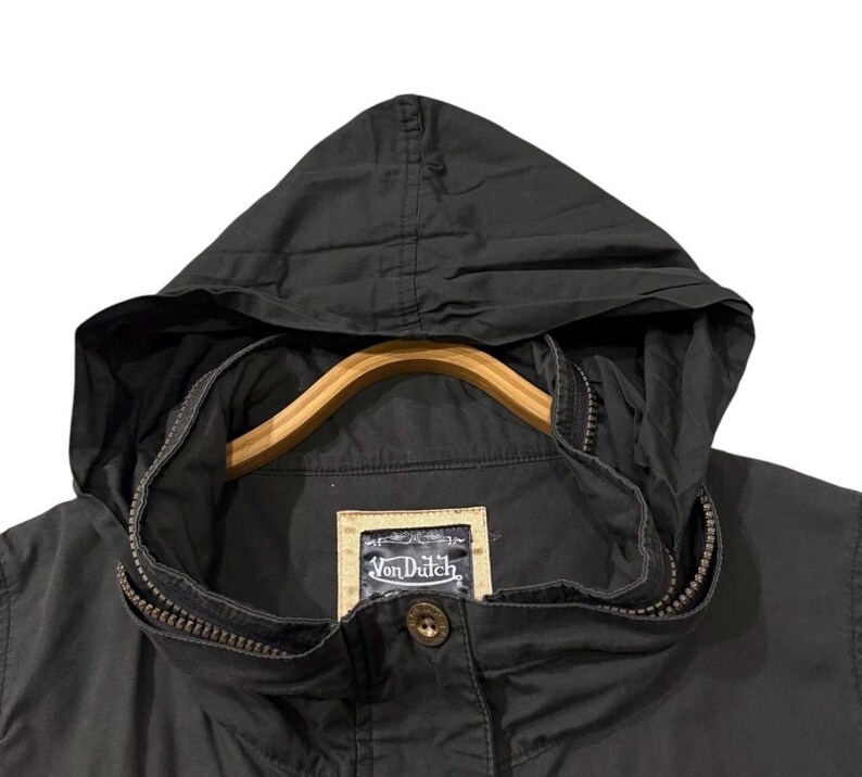 Puede incluir: Chaqueta con capucha negra con cierre de cremallera y percha de madera. La chaqueta presenta una peque&ntilde;a etiqueta con el texto "Von Dutch" y un cierre de bot&oacute;n. La chaqueta es de tela oscura.
