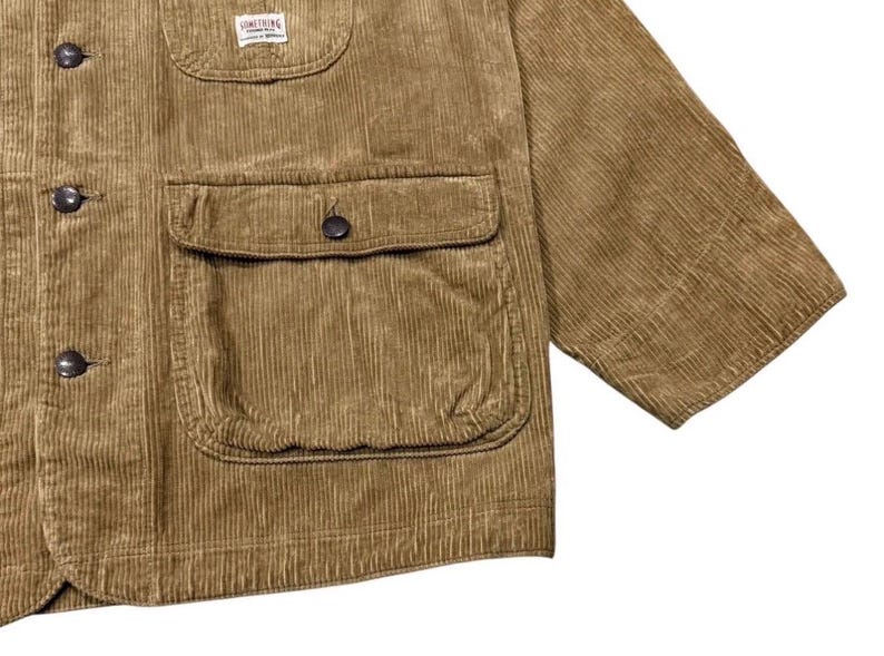 K&ouml;nnte beinhalten: Nahaufnahme einer braunen Cordjacke mit Knopfleiste und zwei gro&szlig;en Vordertaschen. Die Jacke hat ein kleines Etikett auf der oberen Brust und kurze &Auml;rmel. Der Stoff hat eine gerippte Textur.