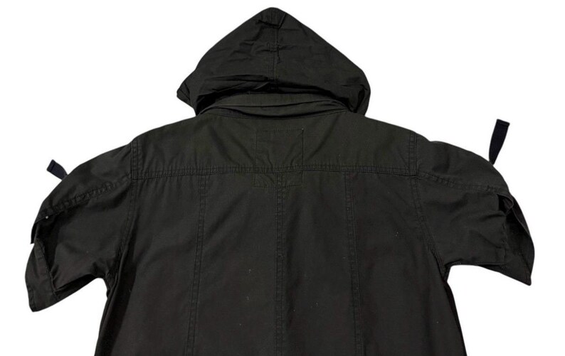 Puede incluir: Chaqueta negra con capucha y mangas cortas. La chaqueta tiene capucha, cierre de botones en la parte delantera y correas ajustables en las mangas. La parte trasera de la chaqueta es visible, mostrando los detalles de las costuras y el dise&ntilde;o general.