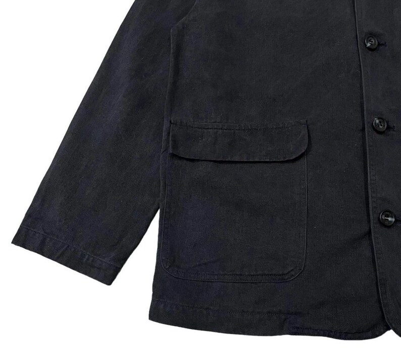 May include: A close-up of a dark navy blue jacket with a single pocket and three buttons. The jacket has long sleeves and a classic design, suitable for various occasions. The fabric appears to be a durable material.