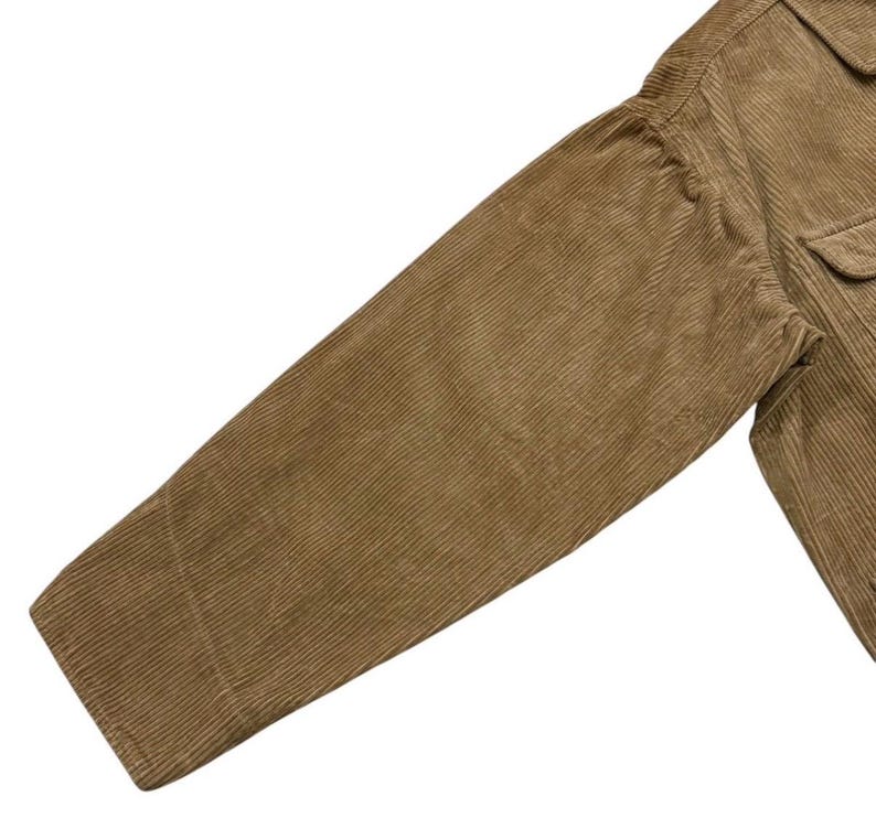 K&ouml;nnte beinhalten: Nahaufnahme eines tan-farbenen Cordjacken&auml;rmels. Der &Auml;rmel ist angewinkelt und zeigt die gerippte Textur des Stoffes. Die Jacke hat eine Tasche und eine gekn&ouml;pfte Manschette. Die Farbe ist ein warmer, erdiger Ton.
