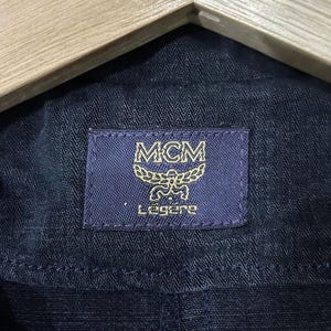 May include: Close-up of navy blue fabric with a rectangular label. The label is dark blue with gold lettering and the MCM logo. The word "L&eacute;g&egrave;re" is printed below the logo.