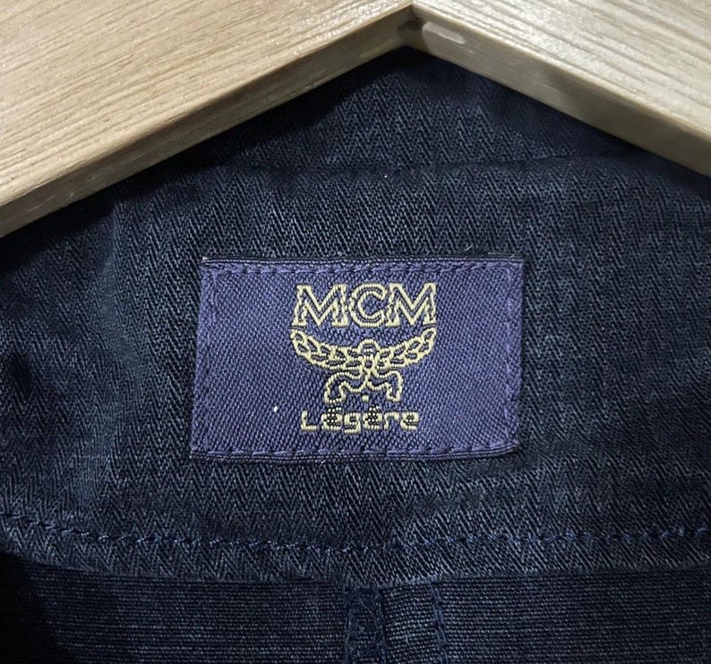 May include: Close-up of navy blue fabric with a rectangular label. The label is dark blue with gold lettering and the MCM logo. The word "L&eacute;g&egrave;re" is printed below the logo.