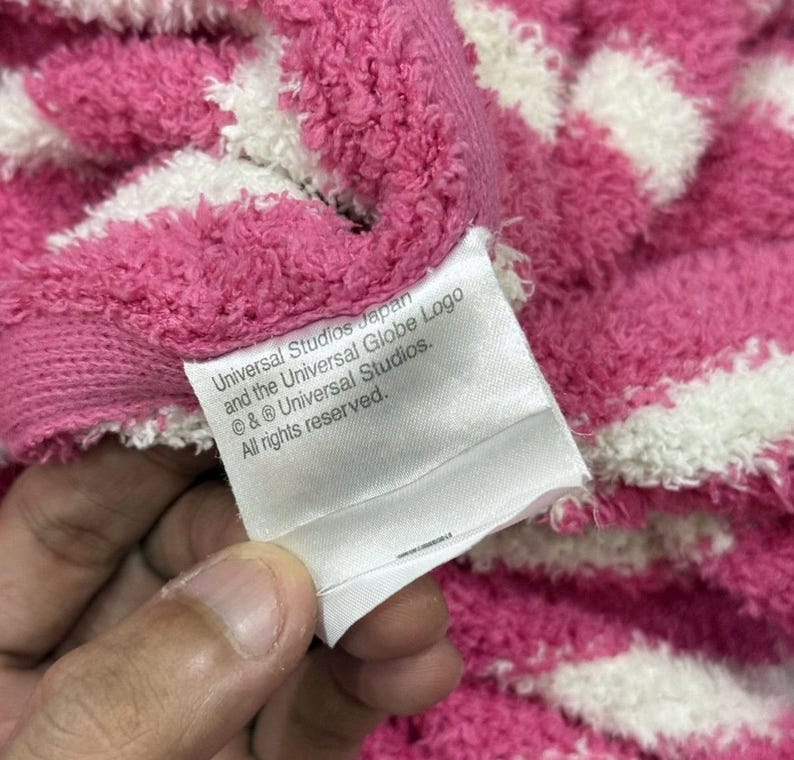May include: Close-up of a pink and white striped towel. A white tag with text that reads "Universal Studios Japan and the Universal Globe Logo &copy; & &reg; Universal Studios. All rights reserved." is visible.