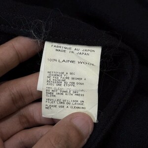 May include: Close-up of a black garment with a white tag. The tag reads "FABRIQUE AU JAPON MADE IN JAPAN 100% LAINE WOOL" and includes care instructions in French and English. The garment appears to be made of wool.