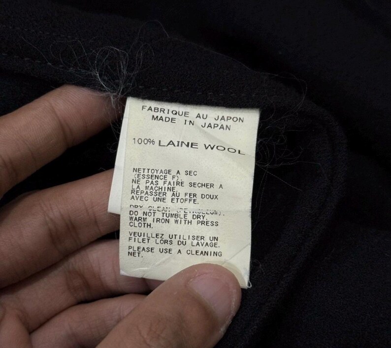 May include: Close-up of a black garment with a white tag. The tag reads "FABRIQUE AU JAPON MADE IN JAPAN 100% LAINE WOOL" and includes care instructions in French and English. The garment appears to be made of wool.