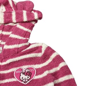 May include: A pink and white striped hooded robe featuring a Hello Kitty patch. The robe has a plush texture and a hood with small, rounded ears. The Hello Kitty patch is heart-shaped with the character's face and the words "Hello Kitty".