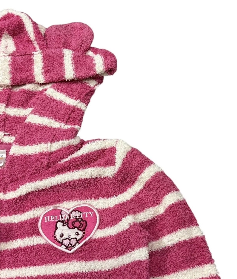 May include: A pink and white striped hooded robe featuring a Hello Kitty patch. The robe has a plush texture and a hood with small, rounded ears. The Hello Kitty patch is heart-shaped with the character's face and the words "Hello Kitty".