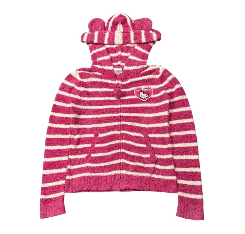 May include: A pink and white striped zip-up hoodie with a hood featuring cat ears. The hoodie has a front pocket and a small emblem on the chest. The design is a cozy, casual style.