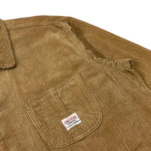 K&ouml;nnte beinhalten: Nahaufnahme einer braunen Cordjacke. Die Jacke hat einen Kragen, eine Brusttasche mit einem kleinen wei&szlig;en Etikett mit der Aufschrift "SOMETHING FOUND 1979" und lange &Auml;rmel. Der Cordstoff hat vertikale Rippen.