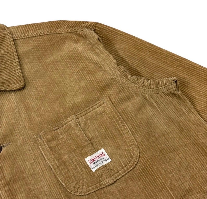 K&ouml;nnte beinhalten: Nahaufnahme einer braunen Cordjacke. Die Jacke hat einen Kragen, eine Brusttasche mit einem kleinen wei&szlig;en Etikett mit der Aufschrift "SOMETHING FOUND 1979" und lange &Auml;rmel. Der Cordstoff hat vertikale Rippen.