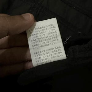 Puede incluir: Primer plano de una etiqueta blanca con texto japon&eacute;s, adjunta a una prenda negra. La etiqueta proporciona instrucciones de cuidado, advirtiendo contra el planchado directo y posibles da&ntilde;os.