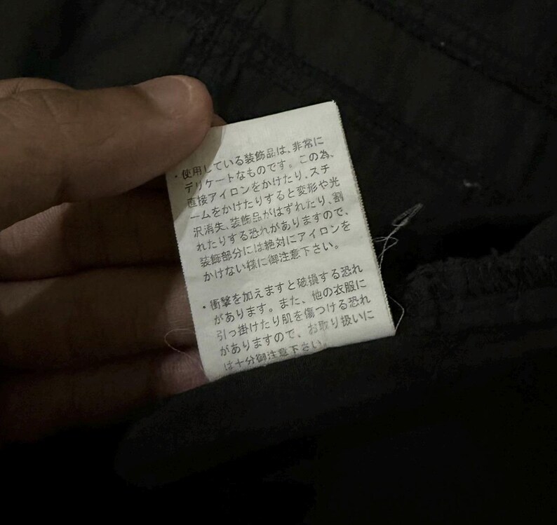 Puede incluir: Primer plano de una etiqueta blanca con texto japon&eacute;s, adjunta a una prenda negra. La etiqueta proporciona instrucciones de cuidado, advirtiendo contra el planchado directo y posibles da&ntilde;os.