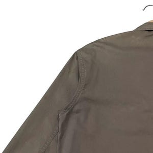 May include: Close-up of a khaki-colored jacket hanging on a metal hanger. The jacket has a collar and a button-down front. The sleeve is visible, and the fabric appears to be a durable material. The jacket is a neutral color, suitable for various outfits.