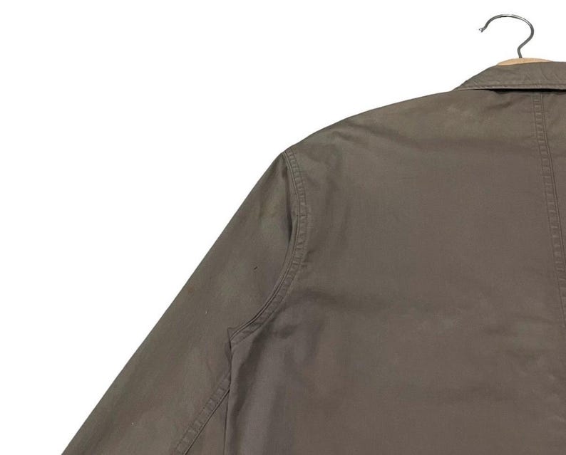 May include: Close-up of a khaki-colored jacket hanging on a metal hanger. The jacket has a collar and a button-down front. The sleeve is visible, and the fabric appears to be a durable material. The jacket is a neutral color, suitable for various outfits.