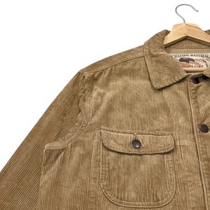 K&ouml;nnte beinhalten: Nahaufnahme einer beigefarbenen Cordjacke mit Knopfleiste und zwei Brusttaschen. Die Jacke hat einen Kragen und ein Etikett mit der Aufschrift "EXCLUSIVE MATERIAL". Die Jacke h&auml;ngt an einem Holzkleiderb&uuml;gel.