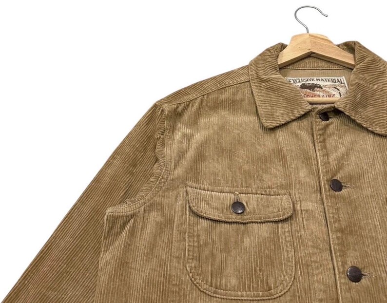 K&ouml;nnte beinhalten: Nahaufnahme einer beigefarbenen Cordjacke mit Knopfleiste und zwei Brusttaschen. Die Jacke hat einen Kragen und ein Etikett mit der Aufschrift "EXCLUSIVE MATERIAL". Die Jacke h&auml;ngt an einem Holzkleiderb&uuml;gel.